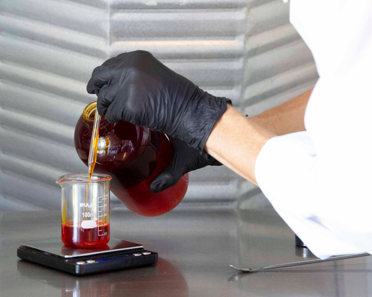 A gloved hand pours a red botanical oil into a glass beaker on a scale