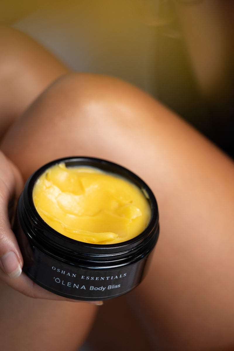 An open jar of ʻŌLENA Body Bliss Balm held against a bare knee