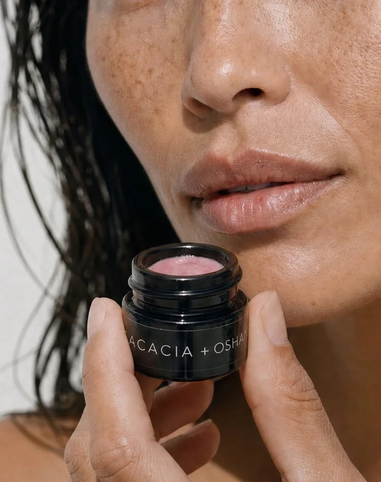 Model with wet hair holding an open jar of OSHAN ACACIA Balm beside her lips.