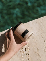 Hand holding the OSHAN CALM ʻĀKALA Body Polish jar on a travertine ledge by the water.
