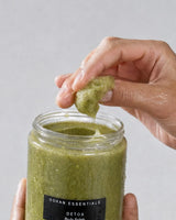 Hand lifting a scoop of OSHAN DETOX ʻŌMAʻOMAʻO Body Polish from the open jar.