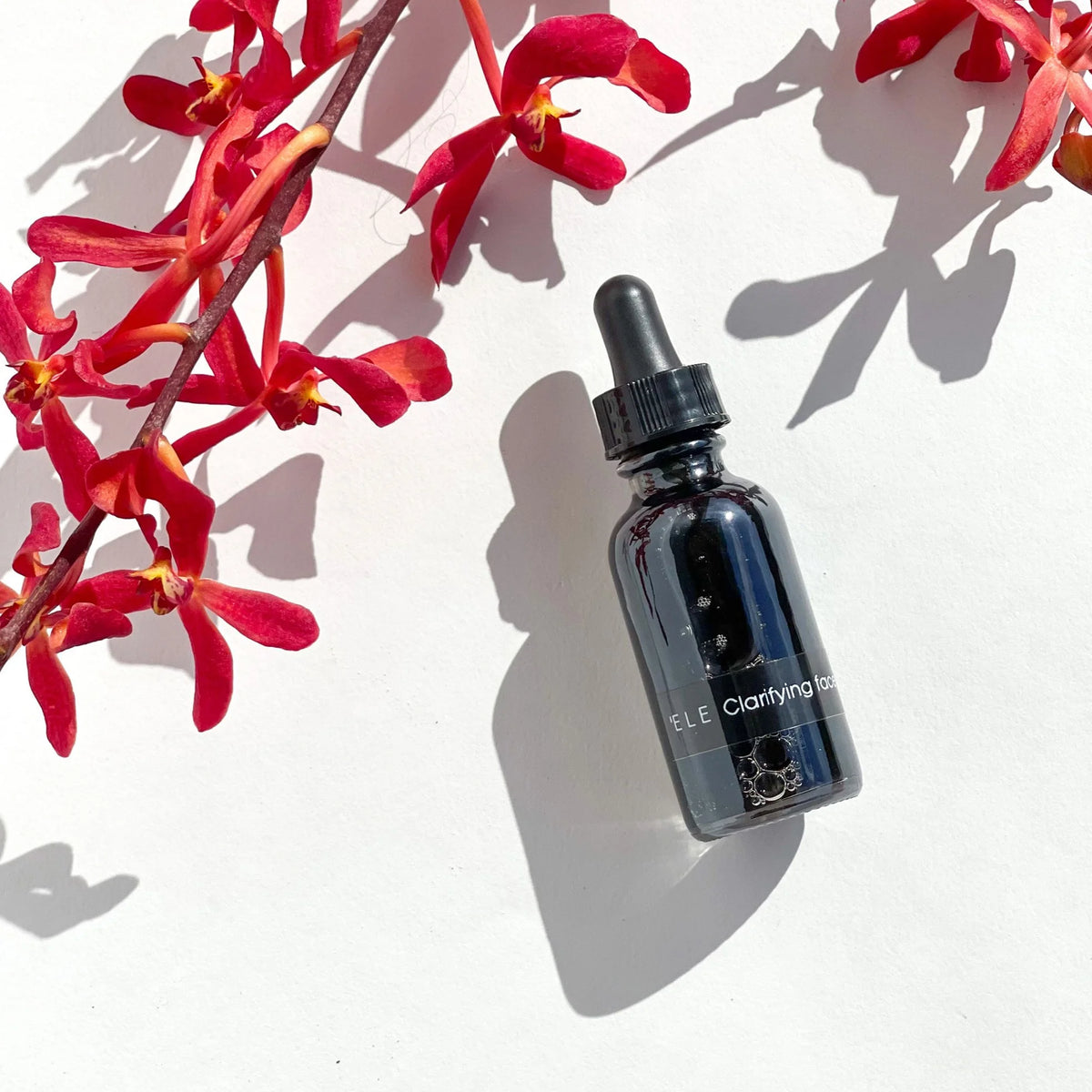 OSHAN ʻELE Clarifying Face Wash bottle among red tropical flowers and leaf shadow.