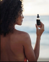 Model at the beach holding a bottle of OSHAN ʻELE Clarifying Face Wash.