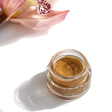 Open jar of OSHAN HULALI Bronzing Face Luminizer beside a pink orchid.