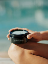Hands holding an open jar of OSHAN KAI After Sun Bliss Balm beside a pool.