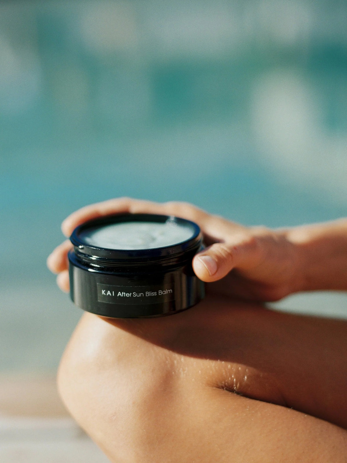 Hands holding an open jar of OSHAN KAI After Sun Bliss Balm beside a pool.