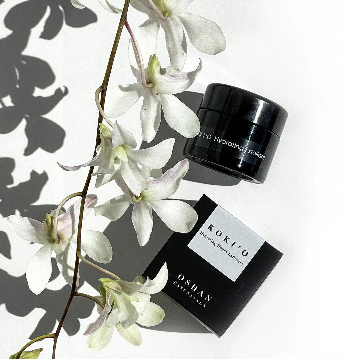 OSHAN KOKIʻO Hydrating Exfoliant jar and box with white orchids and leaf shadow.