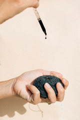 Hand squeezing a wet OSHAN KONJAC Sponge under a dropper of face wash.