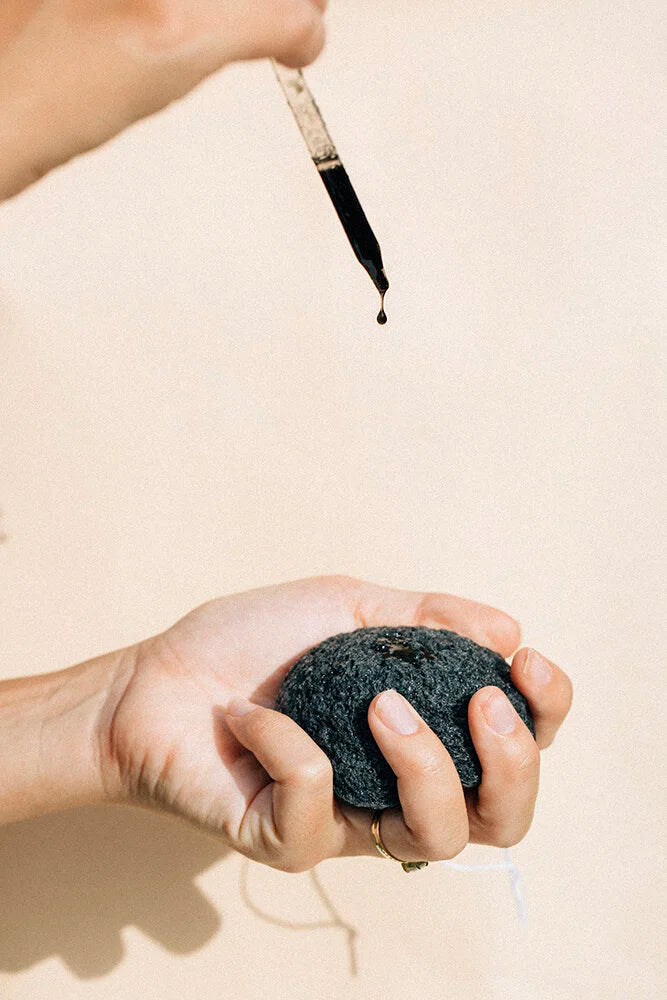 Hand squeezing a wet OSHAN KONJAC Sponge under a dropper of face wash.