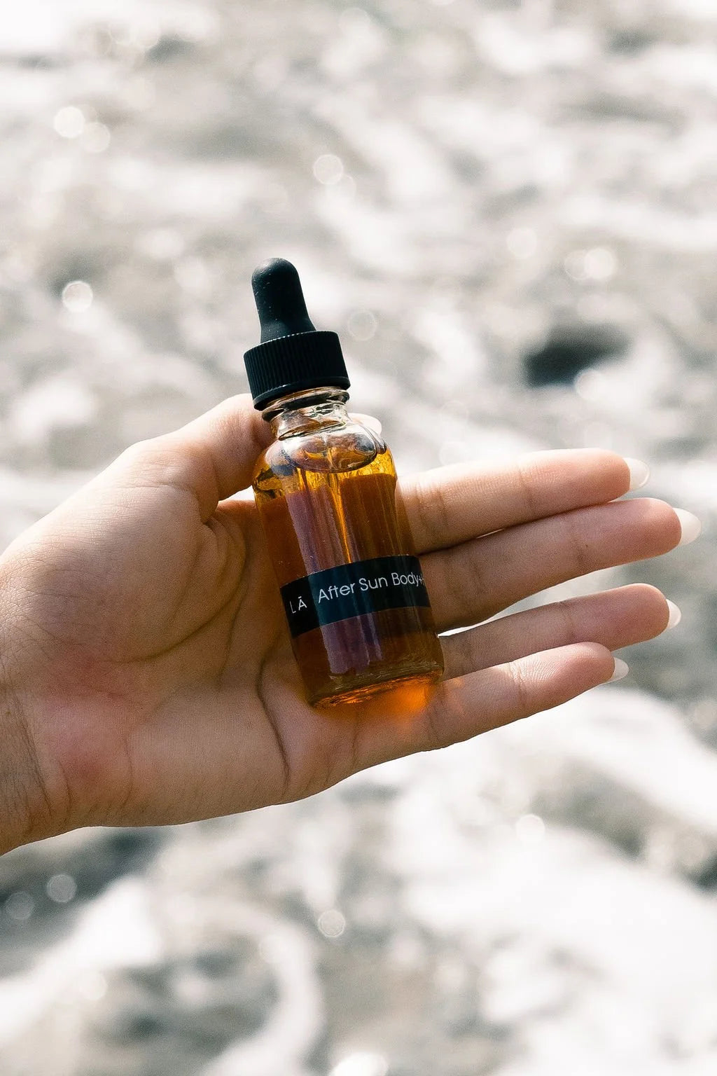 Hand holding the OSHAN LĀ Soothing After Sun Manoʻi bottle above shallow water.