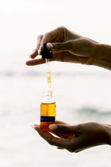 Hands lifting the dropper from an OSHAN LĀ Soothing After Sun Manoʻi bottle at the shore.