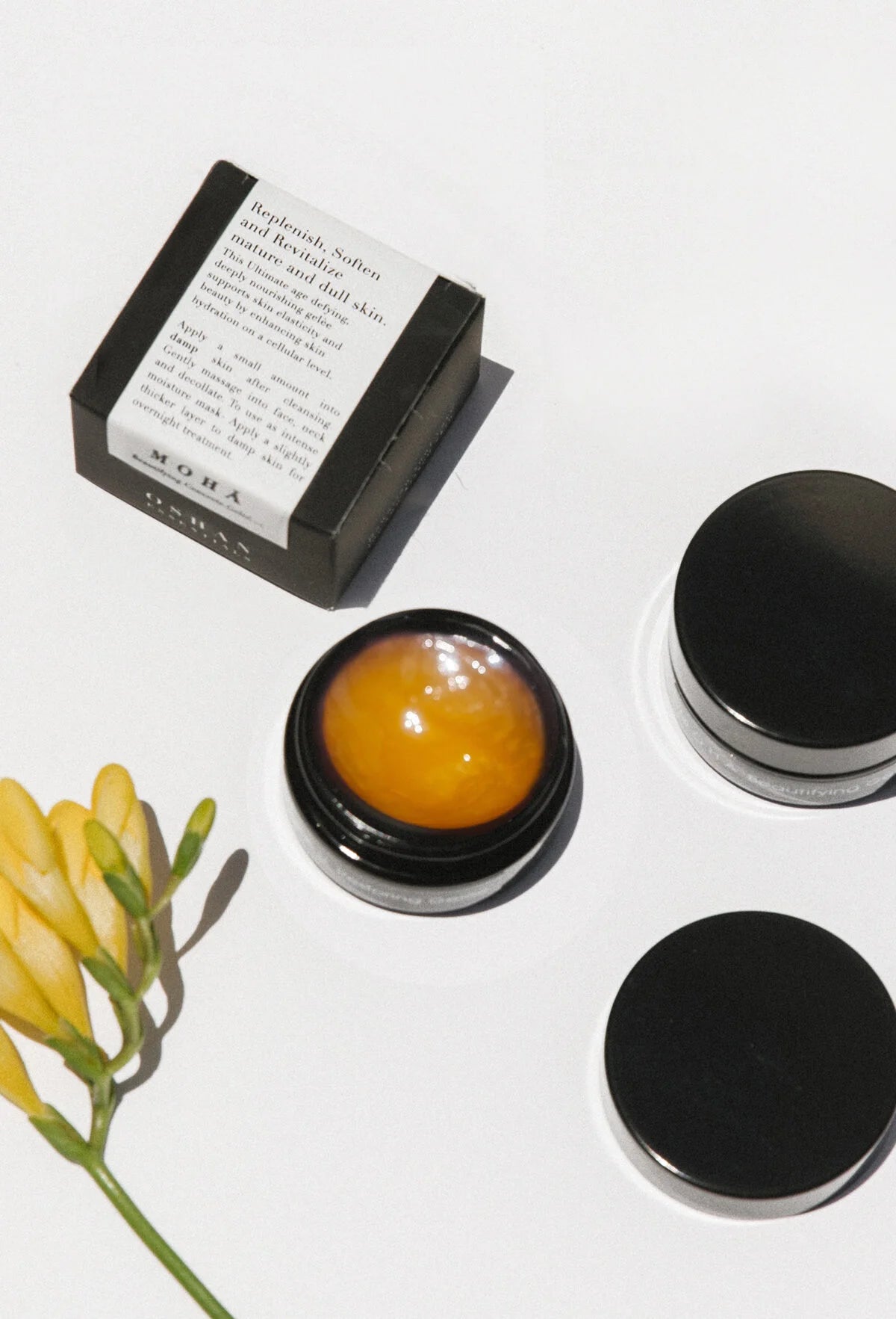 Open jar of OSHAN MOHĀ Beautifying Concrete Gelée with its box and a yellow freesia stem.
