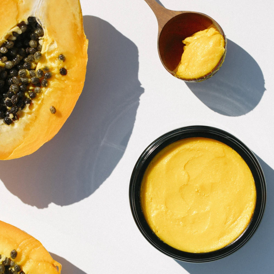 Open jar of OSHAN ʻŌLENA Body Bliss Balm beside a halved papaya and a wooden spoon.