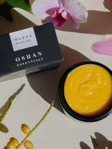 Open jar of OSHAN ʻŌLENA Body Bliss Balm with its box and orchids.
