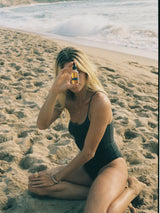 Model sitting on the beach holding the OSHAN WAI Invigorating Hair and Body Manoʻi bottle.