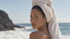 A woman with her hair wrapped in a white towel on a rocky shore by the ocean
